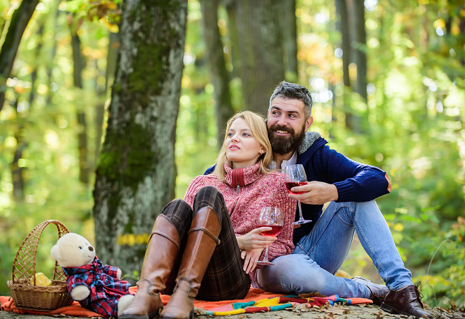 Couple enjoying a cozy autumn picnic in the forest, sitting on a blanket with red wine, surrounded by fallen leaves and soft sunlight