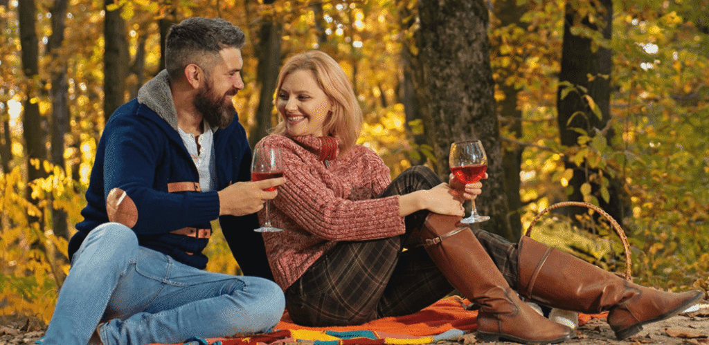 Couple enjoying a romantic autumn picnic in the forest, sitting on a colorful blanket with red wine, surrounded by golden fall foliage.