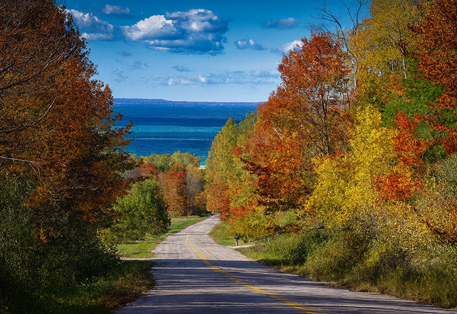 Scenic autumn road winding downhill through colorful forest toward a deep blue lake under a partly cloudy sky.