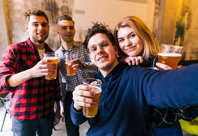 Group of friends taking a selfie indoors, holding beer glasses and smiling in a modern, industrial-style setting.