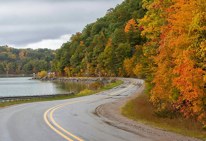 Winding wet road curving along a lake under overcast skies, bordered by vibrant autumn forest with orange, red, and yellow foliage.