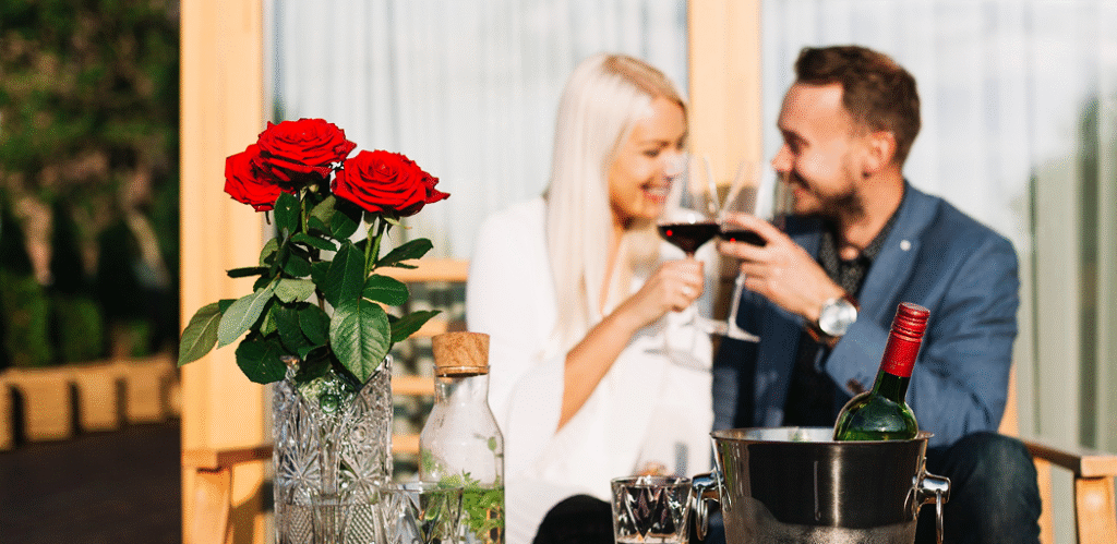 Romantic couple toasting wine on a cozy outdoor patio with red roses, ice bucket, and warm smiles.
