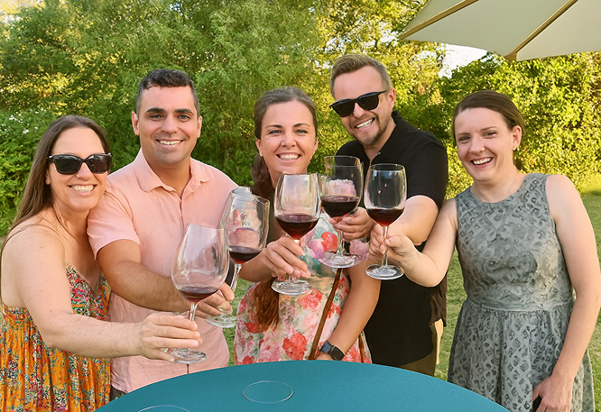 Group of five adults toasting with wine glasses at an outdoor summer gathering, smiling under a garden umbrella with lush greenery in the background.