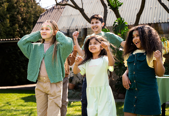 : Five smiling children with face paint enjoying a sunny outdoor celebration on a grassy lawn, with a corrugated-roof building and trees in the background.